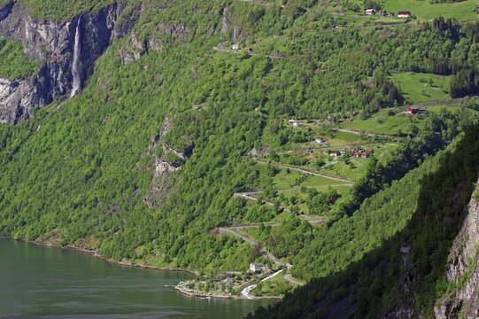 A green, mountainous landscape with a winding road leading up a zig-zag mountain, a few small houses and a waterfall that flows into a river, Eagle Road, Geiranger, Geirangerfjord, Norway