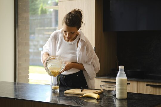 A person blends banana and sea buckthorn in a cozy kitchen. Ingredients are prepared on the counter, with natural light highlighting the freshness of the smoothie-making process