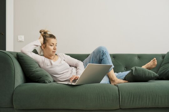 A woman is sitting on a green sofa, scrolling through social media on her laptop. She wears casual clothing and has her hair pulled back