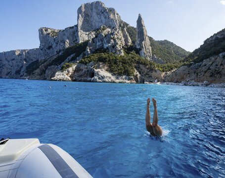 Young woman jumping from a boat into the water, picturesque rocky coast, cliffs with L'Aguglia rock pin, blue sea and Cala Goloritz&eacute; beach, Golfo di Orosei, Baunei, Sardinia, Italy
