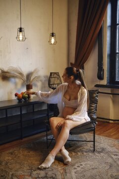 A pregnant woman enjoys her time at home, sitting comfortably in a stylish chair. She reaches for a bowl of fruit placed on a nearby shelf, surrounded by warm lighting and decor