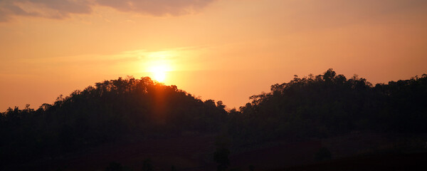 Golden sunset over forested mountain silhouettes beneath a warm orange sky, creating a peaceful natural landscape. Ideal for travel, nature backgrounds, outdoor scenery.