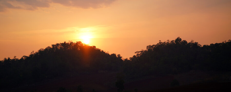 Golden sunset over forested mountain silhouettes beneath a warm orange sky, creating a peaceful natural landscape. Ideal for travel, nature backgrounds, outdoor scenery.
