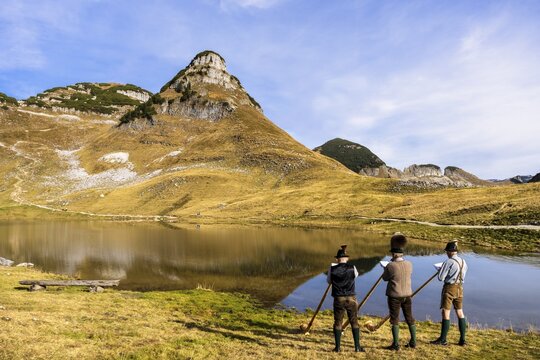 Three men play the alphorn at Lake Augstsee on Mount Loser. The Austrian alphorn trio Klangholz. Mount Atterkogel in the background. Autumn, good weather, blue sky. Reflection. Altaussee, Bad Aussee, Ausseer Land, Totes Gebirge, Styria, Upper Austria, Austria
