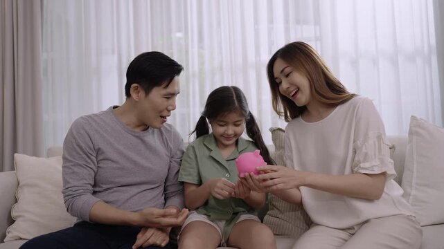 Happy Asian family saving money concept. Smiling father and mother with child girl sitting saving money into a pink piggy bank together for their daughter's future on sofa in the living room at home. 