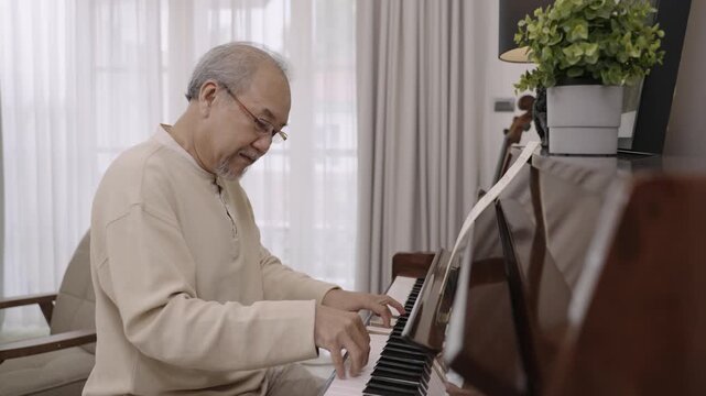 Asian senior man pianist in eyeglasses is sitting playing classical piano alone after retirement for relaxing in living room at home. Elderly male playing a keyboard organ instrument with happiness. 