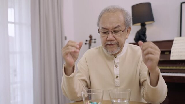 Unhappy Asian senior man sits and looks at pills and antibiotics in a glass with stress in living room at home. Old male needs to take many medicines for his illness. Healthcare for elderly concept. 