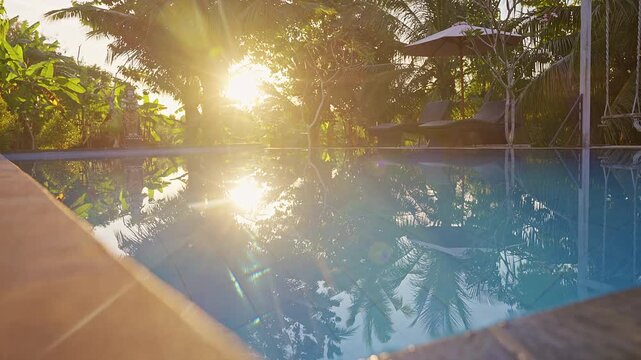 Reflective swimming pool surface illuminated by early morning sunlight in a tropical resort setting. The combination of natural light, palm tree reflections, and calm water creates a tranquil atmosphe