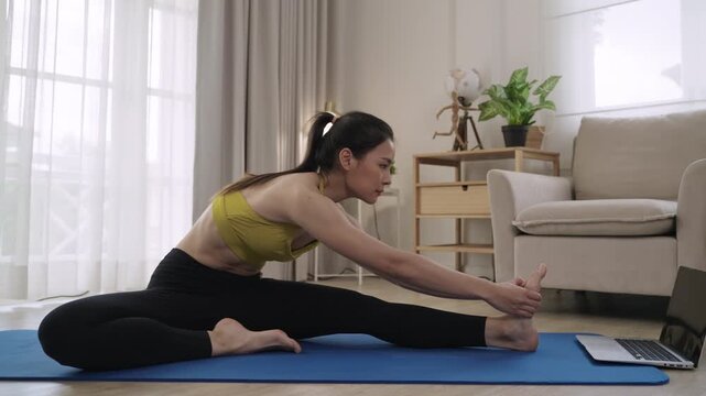 Happy Asian young woman in sportswear watching exercise training online on laptop with exercising on yoga mat for healthcare in living room at home. Slender female practicing a fitness workout class.
