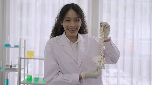 Happy professional woman medical anatomist wearing white lab coat uniform standing holding an anatomical model of the human spine with presentation and explanation carefully detailed in lab at clinic.