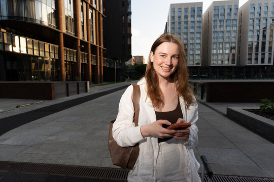 Smiling student pauses with her phone between university buildings