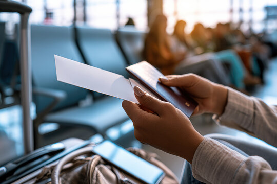 Person holding a boarding pass and passport, preparing for travel in an airport waiting area with chairs and luggage nearby.