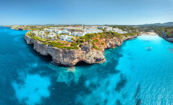 Aerial panorama of sea coast with white resort houses on rocky cliffs. Turquoise blue sea water, sunny sky, boat, sandy beach. Luxury travel destination, Mallorca island, Spain. Drone panoramic view