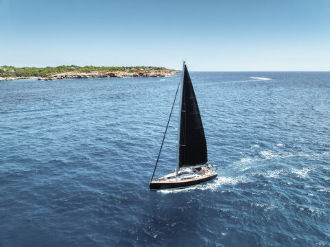 Aerial drone view of luxury sailing yacht with black sails moving in blue sea in summer. Top view of modern sailboat cruising in Mediterranean sea, rocky coastline, Mallorca, Balearic island, Spain