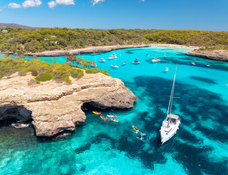 Aerial drone view of yachts and boats anchored in turquoise bay with rocky cliffs, caves, green trees, coastline in Mallorca island, Spain. Clear sea, sunny day. Seascape. Top view. Summer travel