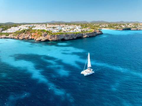 Aerial view of sailing catamaran yacht in turquoise sea water bay. Sailboat. Mallorca, Spain. Luxury boat cruising in ocean cove near sandy beach, resort houses on rocky coast. Drone summer landscape