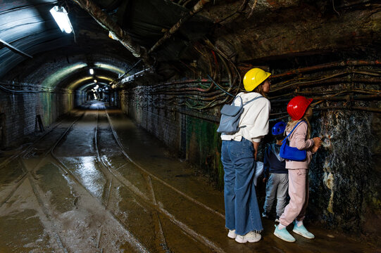 Mother children wear helmet mine tour explores underground mine tunnel, wearing helmets beside wet rail cable rock wall. Curious kids mom discover industrial heritage tourism safety adventure history