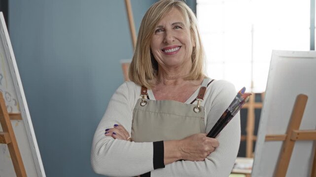 Woman painter holds paintbrushes with crossed arms in studio, smiling with face and hands visible; creative calm contentment.