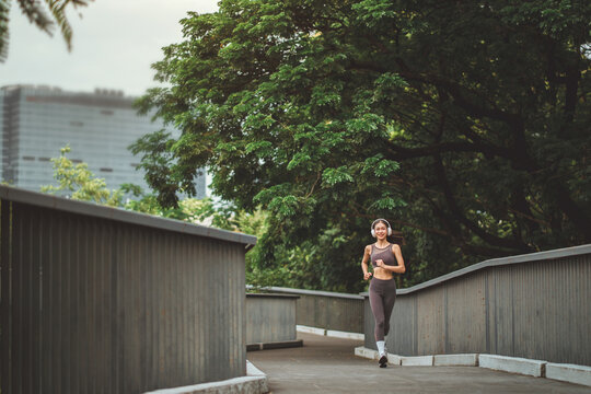 Happy Asian woman in sportswear wearing headphones and running at urban park, cheerful female athlete jogging on bridge pathway surrounded by green trees for fitness, wellness, and active lifestyle