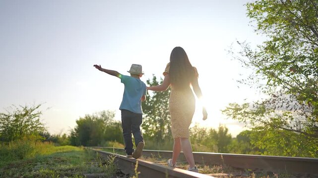 Boy balances on railway track while mother holds his hand at sunset. Child points forward walking on rail with woman supporting. Family explores abandoned railroad in countryside nature.
