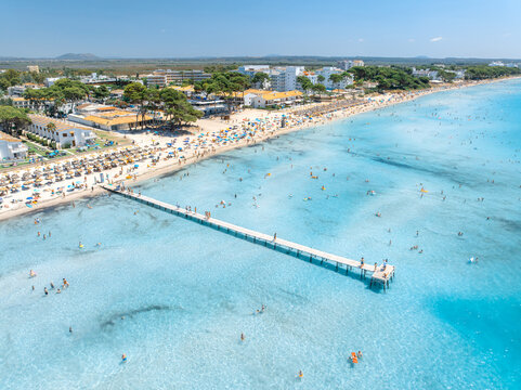 Aerial drone view of Playa de Muro sandy beach in Mallorca, Spain. Wooden pier in turquoise sea water, shore with tourist hotels, mountains, swimming people. Summer vacation. Coastal resort. Top view