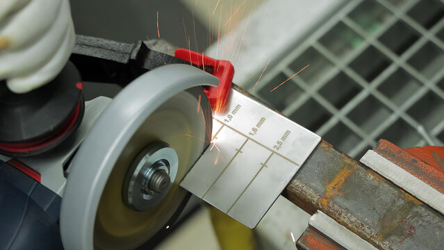 Angle Grinder Cutting Marked Metal Gauge. Close-up angle grinder cuts a marked metal thickness gauge clamped on a rusty workbench, throwing sparks.