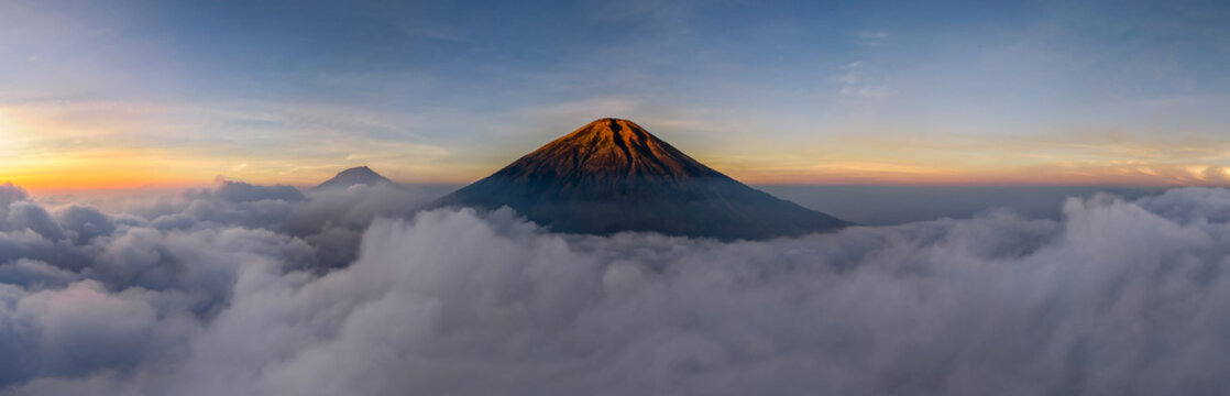 Aerial view of Mount Sindoro and Mount Sumbing rising above a thick sea of clouds during sunrise with golden light illuminating the volcanic peaks in Kecamatan Kedu, Jawa Tengah, Indonesia.