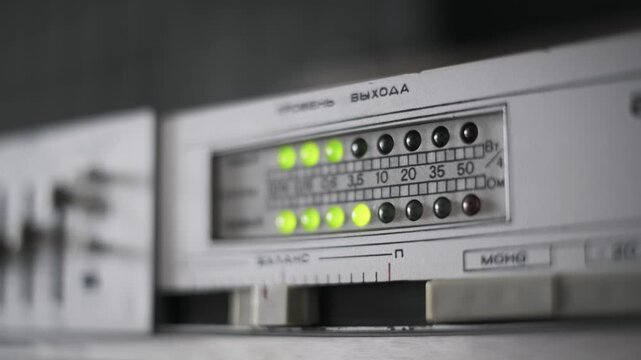 Angled shot with shallow focus on the flashing green LEDs of a Soviet amplifier panel. The fluctuating audio signal with a red peak light suggests dynamic analog sound production.