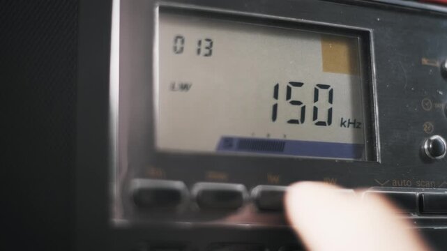 Macro of a finger pressing buttons to switch between radio bands on a digital receiver. The LCD display cycles through FM and SW modes showing MHz and kHz units before tuning.