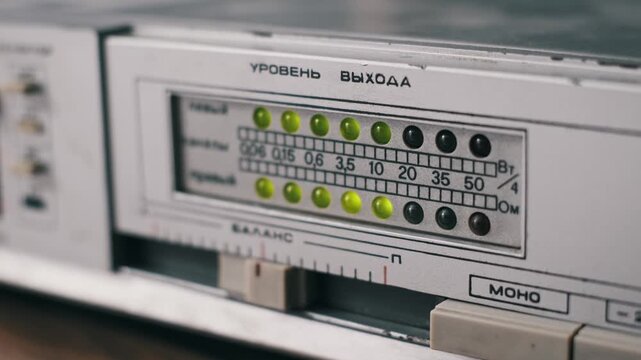 Close-up of a vintage audio amplifier with a blinking LED meter. Yellow-green lights pulse to the music beat with Cyrillic text for Output Level visible on the silver faceplate.