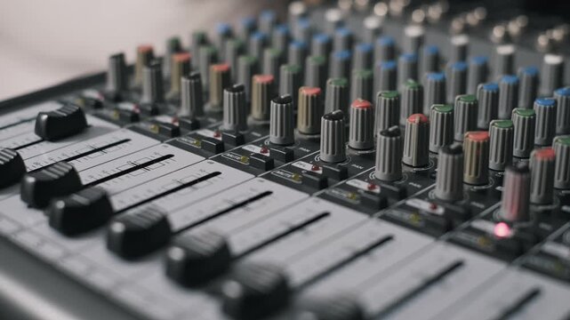 Angled close-up of a multi-channel audio mixing console with a rack focus from foreground faders to colorful EQ knobs. Blinking indicator lights and channel strips are visible in the background.