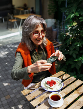 A silver haired woman in red glasses and an orange knit, wrap smiles, she points her phone camera at a strawberry topped plate and a cup of tea.