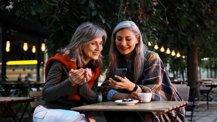 Fototapeta premium A silver haired woman holds a smartphone toward her smiling companion, both are leaning in together at a wooden outdoor table, with coffee and chocolates nearby.