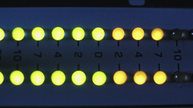 Top-down macro view of stereo VU meter lights flashing on a mixing console. The lights indicate left and right channel audio levels with a numeric decibel scale in a dark studio.