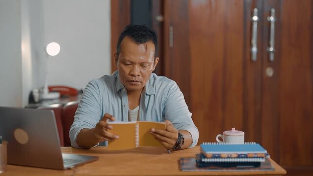 Focus and relaxed of Asian man in casual shirt reading a notebook in home office desk, checking a project on laptop. Learning, analyzing, literacy and education concept.