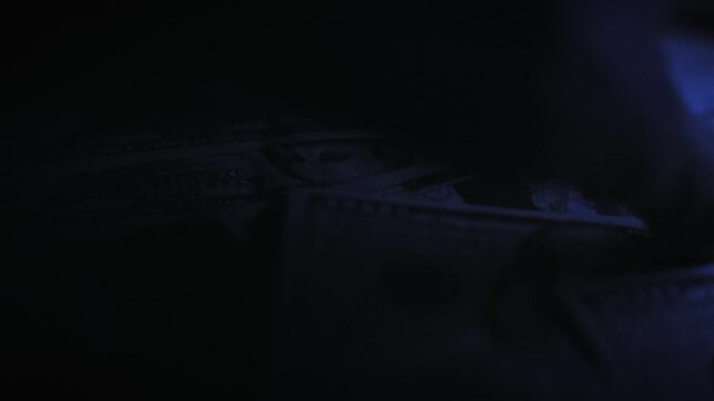 Close-up macro view of hands counting a large stack of one hundred dollar bills under blue light. The rapid movement highlights themes of wealth accumulation and illicit financial transactions.