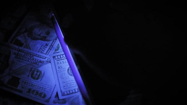 Overhead view of a hand inspecting a disorganized pile of hundred dollar bills with a handheld ultraviolet light. The hidden watermarks and threads glow in the dark for authenticity verification.