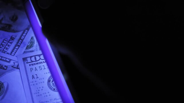 Top-down view of an ultraviolet lamp scanning a scattered pile of hundred dollar bills. The purple light moves across the banknotes to verify authenticity in a dark room.