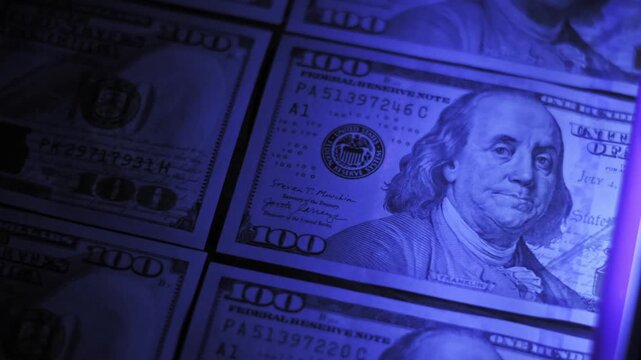 Close-up pan across rows of hundred dollar bills being scanned by a purple ultraviolet light. An expert inspects the banknotes for authenticity and hidden security marks.