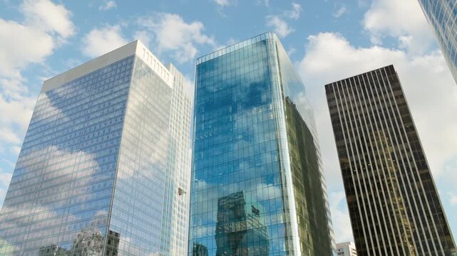 Low-angle panoramic architectural view of modern corporate glass skyscrapers in a bustling financial district, with brilliant reflections of the blue sky and clouds across the multi-story facades.