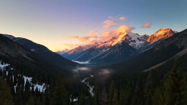 Panoramic alpine landscape featuring snow-capped mountain peaks during sunrise. Golden hour light hits summits above a misty valley with a river, evergreen forests, and silhouetted fir trees.