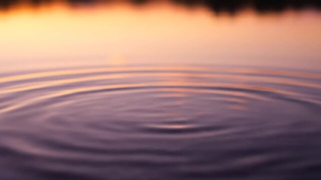 Slow motion macro shot of water drop impact on calm surface creating circular ripples and crown splash with orange sunset reflection bokeh in the background at dusk.
