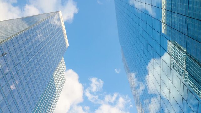 Low-angle architectural view looking up between two modern glass skyscrapers reflecting a clear blue sky and bright white clouds.