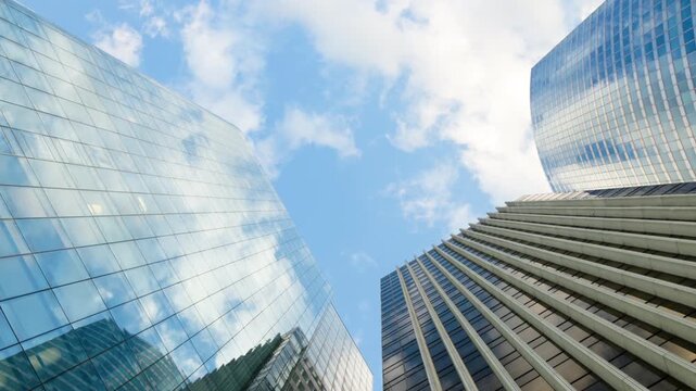 Wide low-angle view looking straight up at corporate skyscrapers with sleek glass and striped facades, capturing the reflection of a bright blue sky and scattered white clouds.