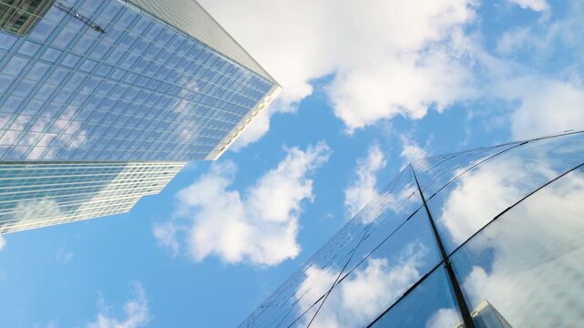 Low-angle architectural view looking straight up into the sky between modern glass skyscrapers, capturing the geometric patterns of the facades and bright reflections of passing clouds.