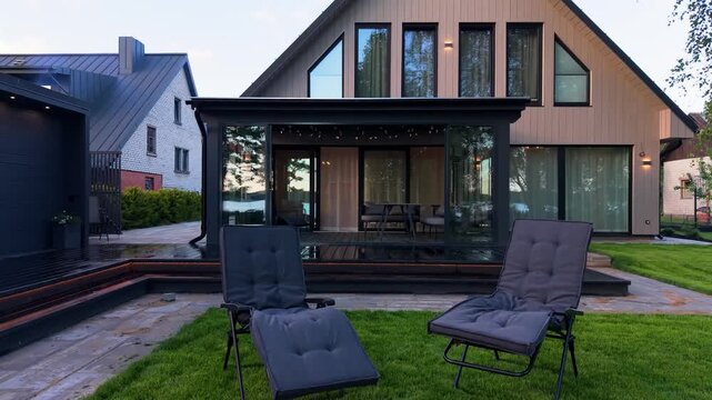 Modern house exterior with glass patio, two lounge chairs, and lush green lawn, showcasing contemporary architecture and outdoor living space