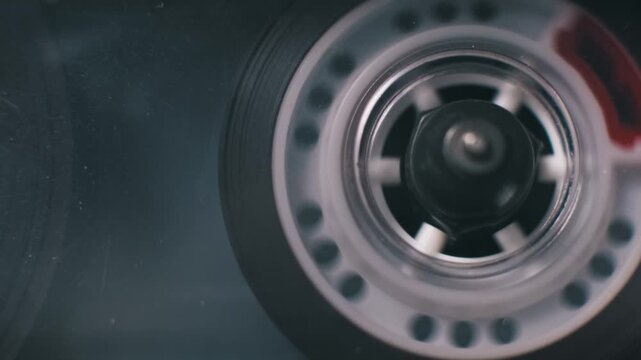 Extreme macro shot of a transparent cassette tape reel spinning inside a vintage player. It reveals a red leader tape section on the spool within the dark, dusty housing.