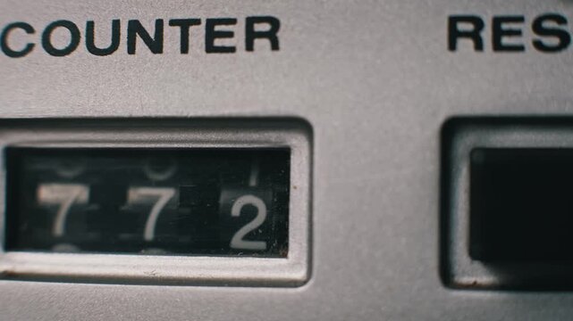 Macro shot of mechanical counter numbers advancing on a vintage silver tape player. Analog digits rotate next to a black reset button to show the current tape position.