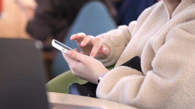Person in cozy beige coat scrolls smartphone. Focus on hands and device. Blurred background suggests casual indoor setting. Suitable for tech, lifestyle, or multitasking themes