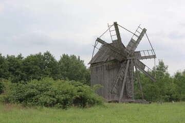 Naklejka premium An old wooden windmill stands tall in a green field under a cloudy sky.